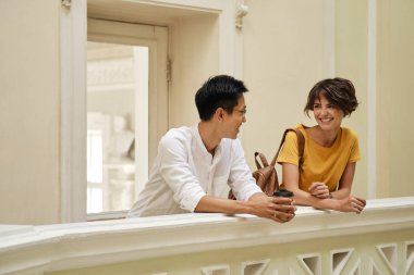 Üniversite koridorunda korkulukya yaslanarak molada sevinçle konuşan iki genç gülümseyen uluslararası öğrenci