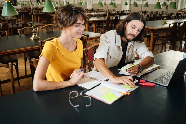 Zwei gut gelaunte junge Studenten machen sich fröhlich Notizen beim gemeinsamen Lernen in der Universitätsbibliothek — Stockfoto