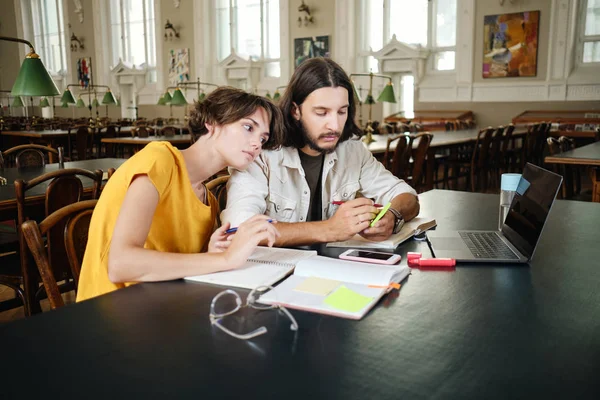 Dois estudantes pensativos jovens que cuidadosamente fazem notas que estudam com o computador portátil em conjunto na biblioteca da universidade — Fotografia de Stock