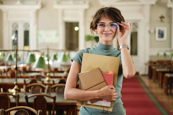 Ritratto di bella studentessa in occhiali con taccuini che guarda felicemente in macchina fotografica nella biblioteca dell'università — Foto Stock