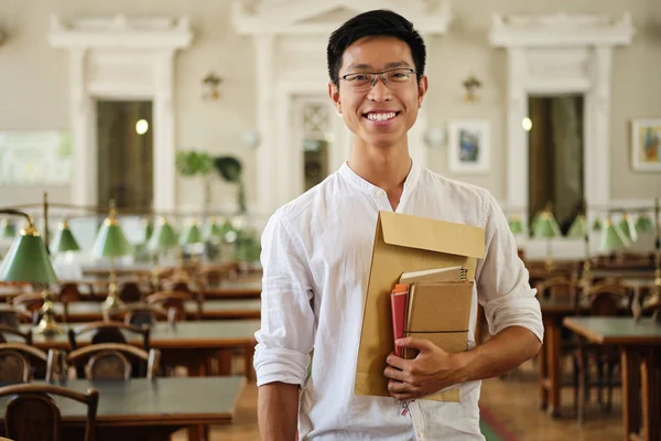 Молодий усміхнений азіатський студентка-чоловік в окулярах з блокнотами радісно дивиться в камеру в бібліотеці університету — стокове фото