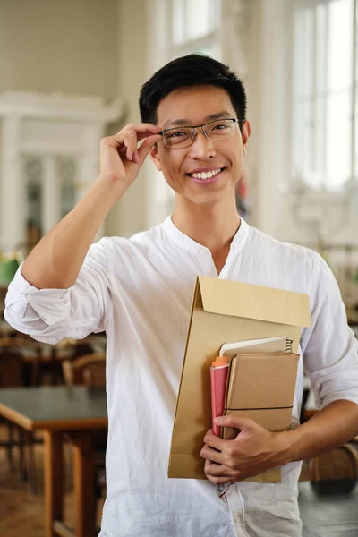 Молодий привабливий азіатський студентка-чоловік в окулярах з книгами радісно дивиться в камеру в бібліотеці університету — стокове фото