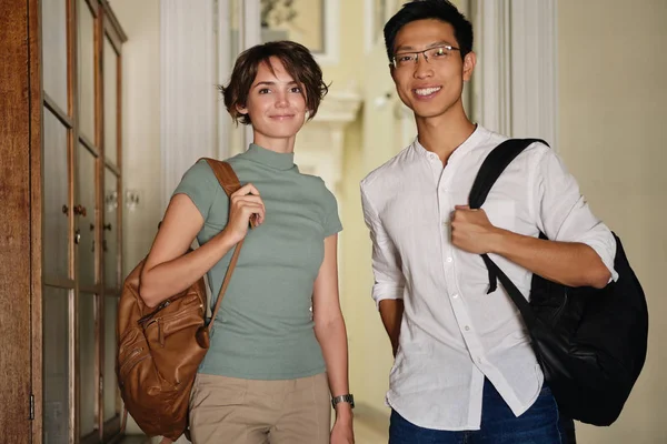 Two young attractive international students with backpacks happily looking in camera standing in corridor of university