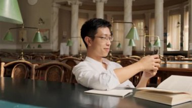 Üniversite kütüphanesinde mutlu cep telefonu kullanarak genç Asya erkek öğrencinin çekim takibi