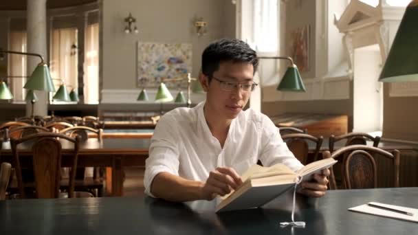 大学の図書館でカメラの読書本を楽しそうに見ているアジアの若い男子学生のショットで追跡 — ストック動画