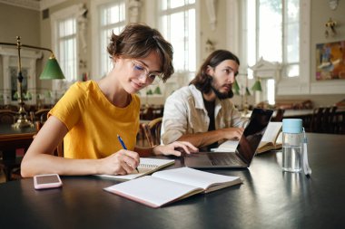 Genç çekici gülümseyen kız öğrenci mutlu üniversite kütüphanesinde dizüstü bilgisayar ile çalışma notları yazma