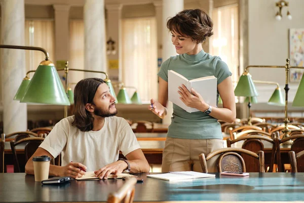 Giovane bella tutor femminile con libro che spiega allo studente nuovo tema durante la lezione in biblioteca dell'università — Foto Stock