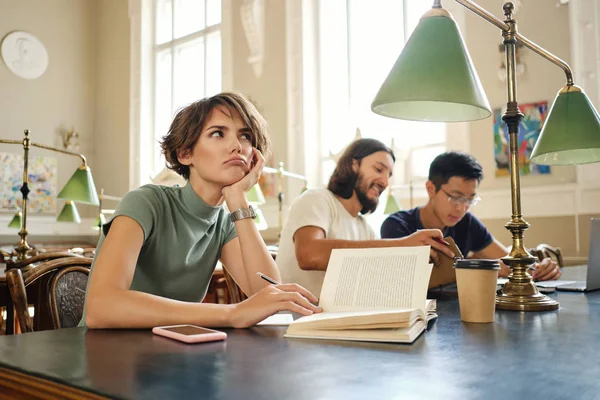 Ung kvinnlig student med bok eftertänksamt arbetar med nytt studieprojekt i Universitetsbiblioteket — Stockfoto