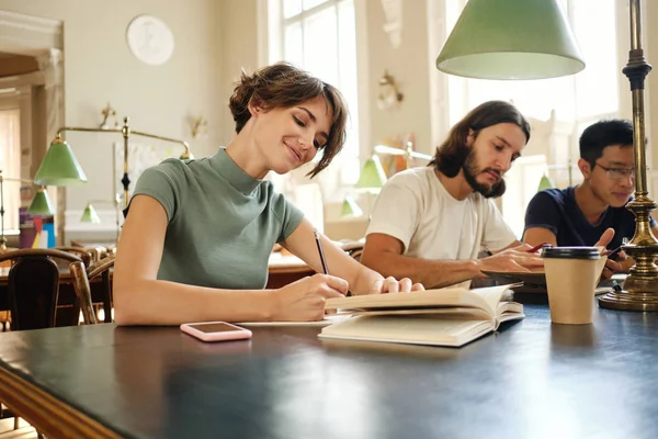 Młoda atrakcyjna studentka z książką szczęśliwie pracuje nad nowym projektem studiów w bibliotece Uniwersytetu — Zdjęcie stockowe