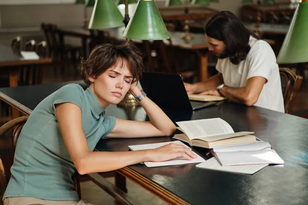 Genç yorgun kız öğrenci yorgun üniversite kütüphanesinde eğitim sırasında kitaplarla masa üzerinde uyku — Stok fotoğraf