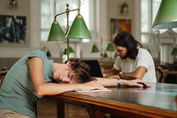 Молодая уставшая студентка спит на столе во время учебы в библиотеке университета — стоковое фото