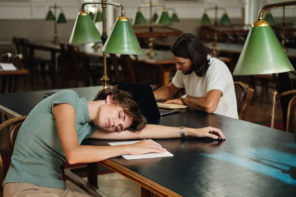 Молода втомлена студентка з блокнотом спить на столі під час навчання в бібліотеці університету — стокове фото