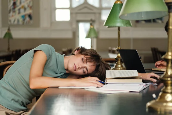 Ung sorglig trött kvinnlig student sova på skrivbord med böcker runt under studie i biblioteket av universitet — Stockfoto