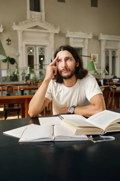 Unga vemodigt manliga studerande eftertänksamt studera ensam med böcker i biblioteket på universitetet — Stockfoto