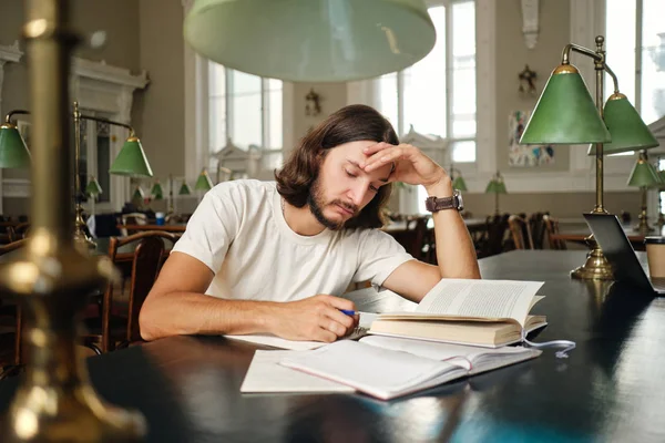 Unga vemodigt manliga studenten omtänksamt skriva anteckningar förbereder sig för examen i biblioteket för universitet — Stockfoto