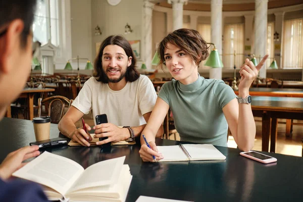 Junge attraktive Gelegenheitsstudenten diskutieren in Bibliothek der Universität fröhlich über Studienprojekt — Stockfoto