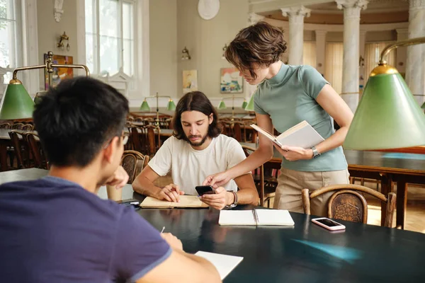 Giovane tutor femminile con libro aiutare gli studenti con lo studio durante la lezione in biblioteca dell'università — Foto Stock