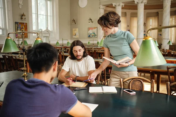 Giovane tutor femminile con libro che spiega agli studenti il nuovo tema durante la lezione in biblioteca dell'università — Foto Stock