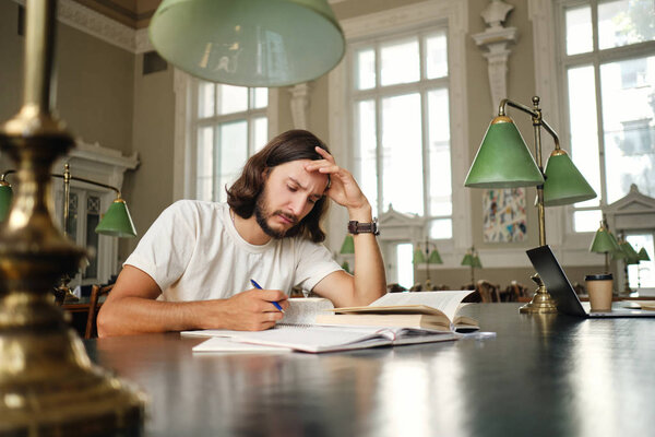 Young tired male student thoughtfully preparing to exam with book and study notes in library of university Stock Photo