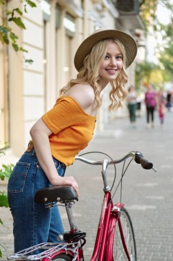 Şehir caddesinde klasik bisiklete yaslanmış kameraya mutlu bir şekilde bakan şapkalı çekici gülümseyen sarışın kız