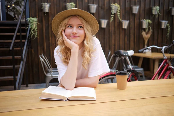 Gyönyörű romantikus szőke lány kalapban támaszkodva a kezét, és álmodik a könyv és kávé udvarán kávézó — Stock Fotó