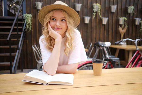 Красивая блондинка в шляпе, мечтательно оглядывающаяся с книгой и кофе на столе во дворе кафе — стоковое фото