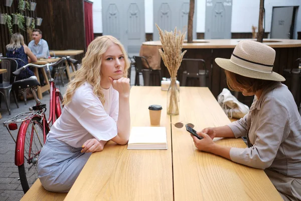 Chica rubia seria agresivamente mirando en la cámara mientras su amigo cerca de usar el teléfono celular en el patio de la cafetería — Foto de Stock