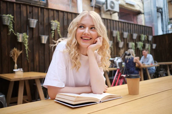 Pretty cheerful casual blond girl happily looking away with book and coffee on table in courtyard of cafe — Stock Photo, Image