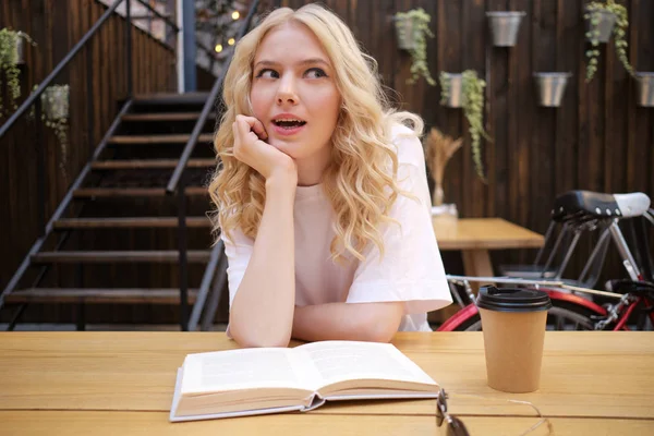 Attractive astonish blond girl leaning on hand amazedly looking away with book and coffee in courtyard of cafe — Stock Photo, Image