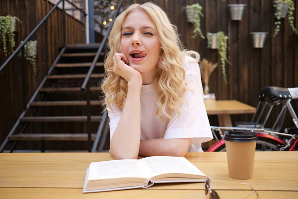 Attractive blond girl playfully showing tongue in camera sitting with book and coffee in courtyard of cafe — Stock Photo, Image