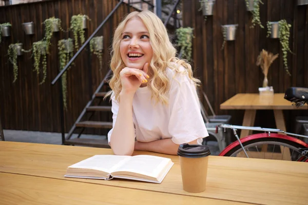 Beautiful smiling blond girl happily dreaming sitting with book and coffee in courtyard of cafe — Stock Photo, Image