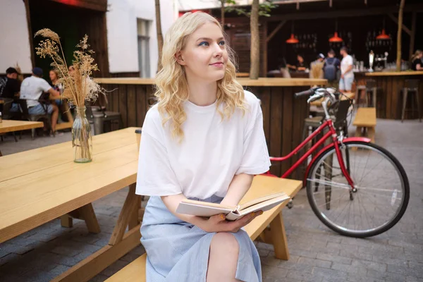 Beautiful romantic blond girl thoughtfully reading book resting alone in courtyard of cafe — Stock Photo, Image