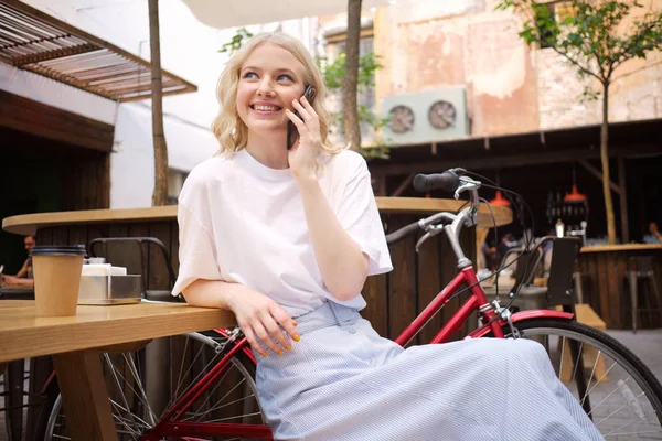 Attractive cheerful blond girl happily talking on cellphone resting in courtyard of cafe — Stock Photo, Image