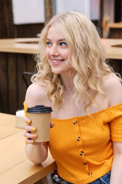 Привлекательная улыбающаяся блондинка счастливо смотрит прочь с кофе, чтобы пойти в руку во дворе кафе — стоковое фото
