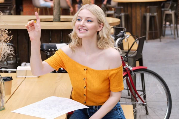 Красивая улыбающаяся блондинка счастливо делает заказ сидя с меню во дворе кафе — стоковое фото