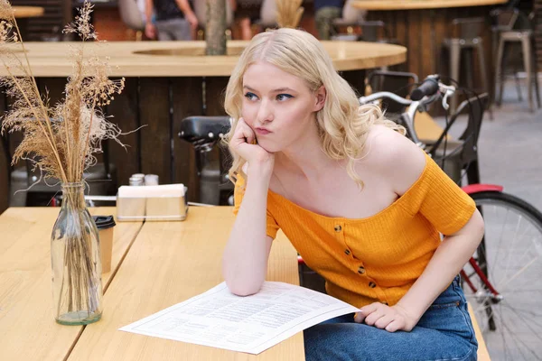 Attractive blond girl thoughtfully deciding what to order sitting with menu in courtyard of cafe — Stock Photo, Image