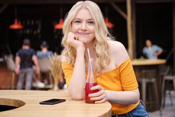 Портрет симпатичной улыбающейся блондинки, счастливо смотрящей в камеру и пьющей лимонад во дворе кафе — стоковое фото