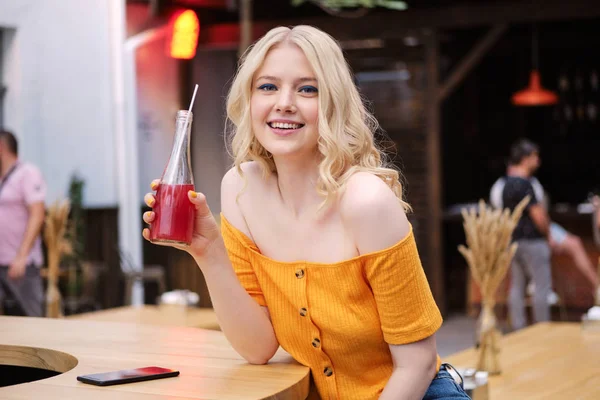 Красивая улыбающаяся блондинка счастливо смотрит в камеру, а лимонад отдыхает во дворе кафе — стоковое фото