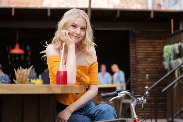 Красивая романтическая блондинка, опирающаяся на руку мечтательно глядя в камеру с лимонадом во дворе кафе — стоковое фото