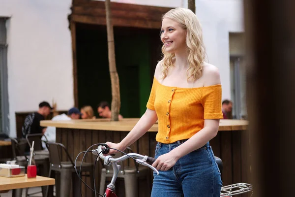 Vista lateral de chica rubia bastante sonriente alegremente de pie con bicicleta clásica en el patio de la cafetería — Foto de Stock