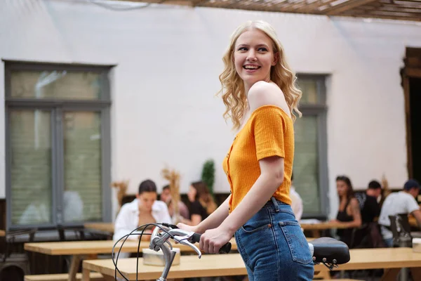 Приваблива усміхнена блондинка радісно дивиться в сторону стоячи з класичним велосипедом у дворі кафе — стокове фото