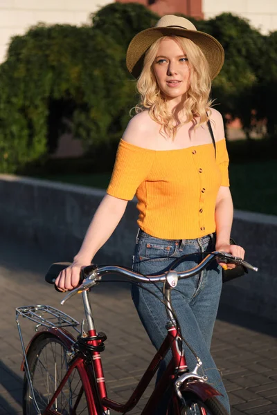 Beautiful casual blond girl in hat dreamily walking with classic bicycle on city street — Stock Photo, Image