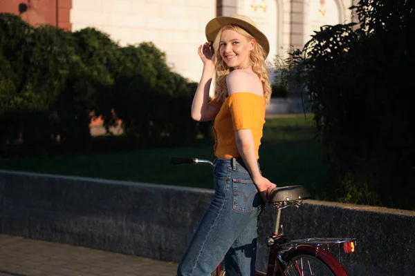 Hermosa chica rubia romántica en sombrero de pie con bicicleta mirando alegremente en la cámara en la calle de la ciudad — Foto de Stock