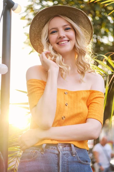 Retrato de menina loira sorridente atraente em chapéu olhando alegremente na câmera sobre o pôr do sol na rua da cidade — Fotografia de Stock