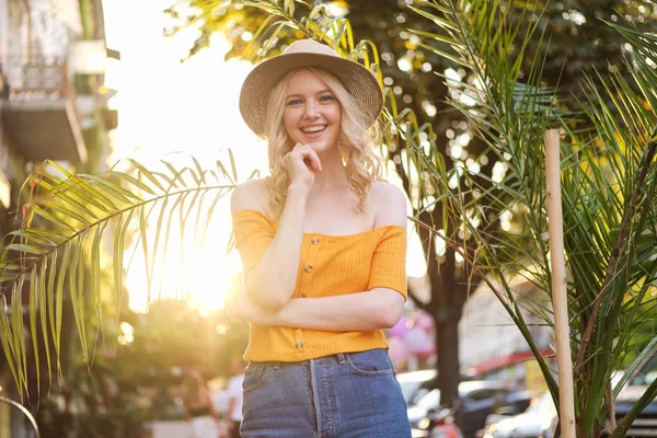 Beautiful romantic blond girl in hat joyfully looking in camera over sunset on city street — Stock Photo, Image