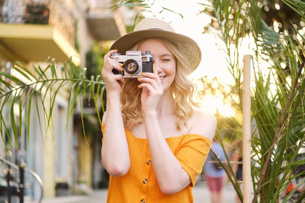 Pretty smiling blond girl in hat happily taking photo on retro camera on city street — Stock Photo, Image