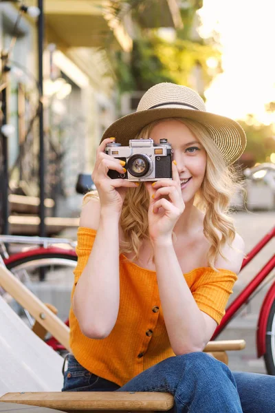 Довольно обычная блондинка в шляпе мечтательно фотографирует на ретро-камеру на шезлонге в уличном кафе — стоковое фото