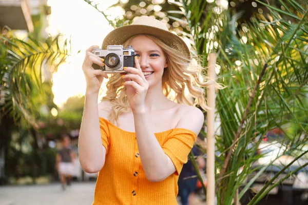 Atractiva encantadora chica rubia sonriente en sombrero felizmente tomar fotos en la cámara retro en la calle de la ciudad —  Fotos de Stock