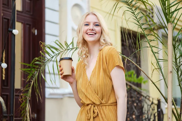 Portrait of beautiful cheerful blond girl in dress with coffee to go joyfully looking in camera on city street — Stock Photo, Image