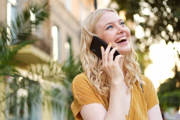 Portrait of pretty cheerful blond girl joyfully looking away talking on cellphone on city street — Stok Foto